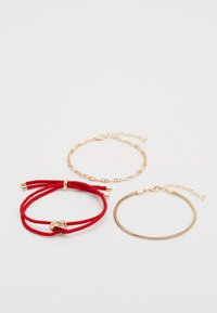 Trois bracelets : un bracelet en corde rouge avec un accent doré, un bracelet en chaîne dorée et un bracelet en fil doré flexible avec un fermoir.