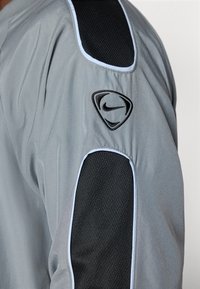 Nike-jakke med gråt stof, der har et sort net-panel på skulderen, lyseblå detaljer og et broderet Nike-logo på ærmet.
