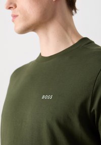 Olivově zelené tričko vyrobené z bavlny s kulatým výstřihem; na hrudi má decentní logo "BOSS" ve světle šedé barvě. Měkká textura.