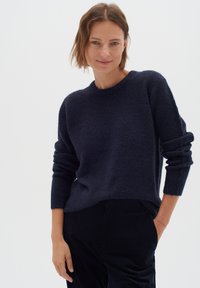 Tumesinine kootud sviiter pehme, tekstuurse materjali ja ümmarguse kaelusega, pikkade varrukatega. Kombineeritud mustade pükstega, stiililt juhuslik.