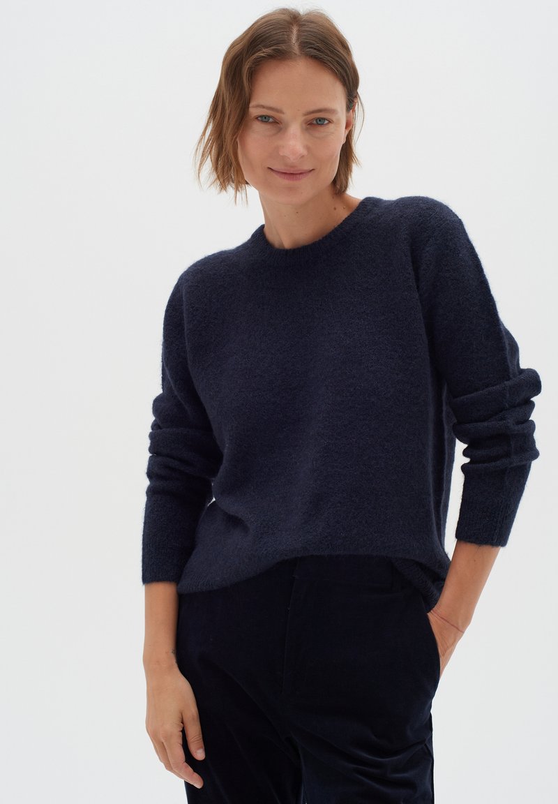 Tumesinine kootud sviiter pehme, tekstuurse materjali ja ümmarguse kaelusega, pikkade varrukatega. Kombineeritud mustade pükstega, stiililt juhuslik.