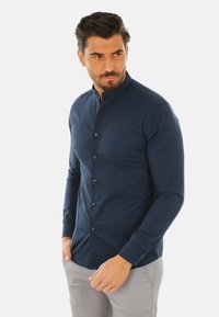 Camicia a maniche lunghe blu navy con colletto alla cinese, chiusura con bottoni e texture liscia, abbinata a pantaloni grigi chiari.