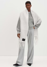 Sciarpa di lana bianca con frange, abbinata a un maglione grigio texture e pantaloni larghi grigio chiaro. Scarpa nera con decorazioni.