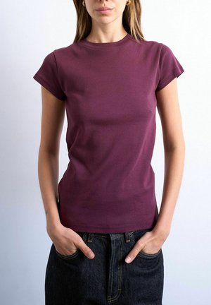 Woman wearing a fitted short-sleeve purple top with hands in dark denim jeans pockets against a plain background.