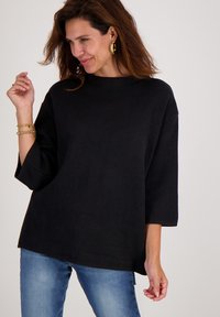 Vrouw met bruin haar, gekleed in een losse zwarte trui, blauwe jeans, gouden oorringen en armbanden, poserend tegen een effen lichte achtergrond.