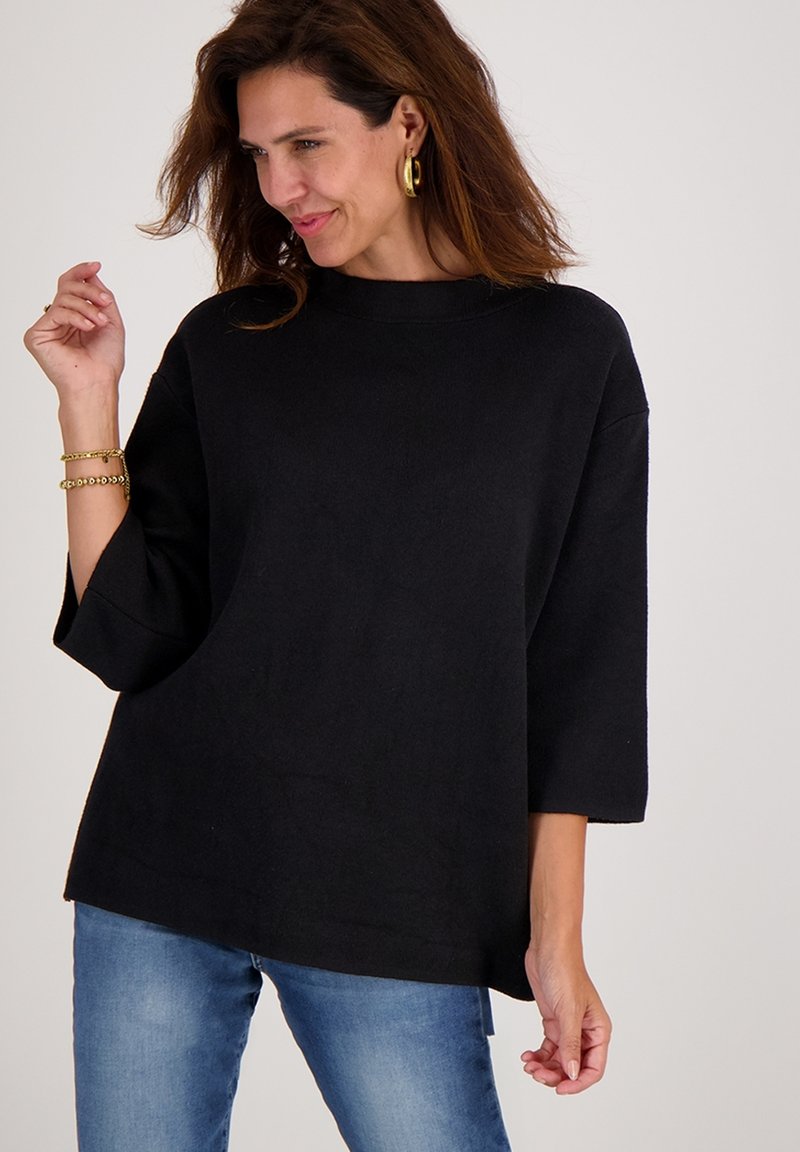 Vrouw met bruin haar, gekleed in een losse zwarte trui, blauwe jeans, gouden oorringen en armbanden, poserend tegen een effen lichte achtergrond.