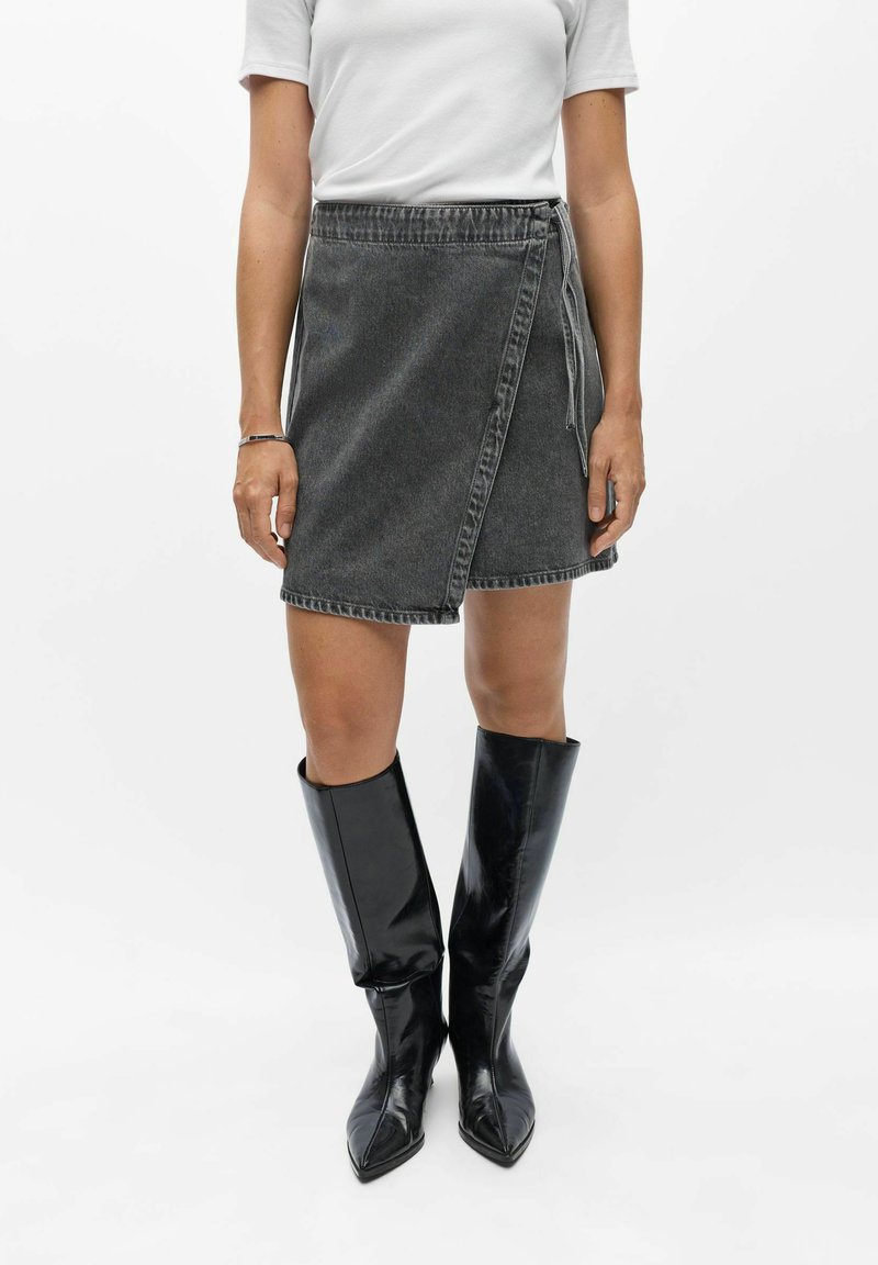 Femme portant une chemise blanche à manches courtes, une jupe en jean noire portefeuille et des bottes noires brillantes jusqu'aux genoux, debout devant un fond blanc.