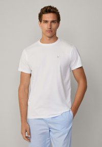 Camiseta blanca de manga corta hecha de una tela suave. Presenta un escote redondo y un pequeño logo gris en el lado izquierdo del pecho.