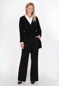 Zwarte gebreide wikkelcardigan met een riem in de taille, versierd met een textuurpatroon, gedragen over een witte blouse met kraag en zwarte wijde broek.