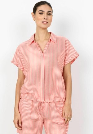 Frau trägt ein kurzärmeliges Hemd mit hellrosa und roten vertikalen Streifen und passende Hosen mit Kordelzug, steht vor einem weißen Hintergrund.