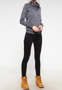 Sweat-shirt gris à col châle avec de petits motifs multicolores, associé à des leggings noirs et des bottes lacées couleur beige, présentant un tissu texturé.