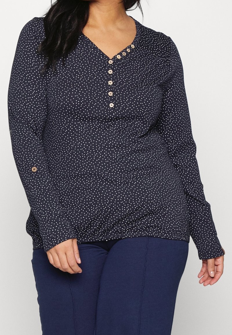 Långärmad marinblå topp med vita prickar, V-ringning och knappdetaljer vid halsen och ärmarna. Mjuk, texturerad tyg.