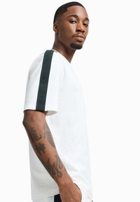 Witte T-shirt met korte mouwen, voorzien van een zwarte zijstreep met reliëftekst. De stof lijkt zacht en het heeft een relaxte pasvorm.