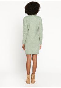 Robe pull tricotée en vert pâle avec un col montant, des poignets côtelés et des détails en cables complexes sur les manches. Associée à des bottines beiges.