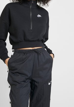 Person iført en sort Nike cropped zip-up sweatshirt og sorte snøreløserebukser med hvide trykknapper på siderne og Nike-logo.