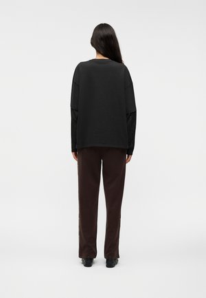 Personne aux longs cheveux foncés, debout de dos, portant une chemise noire oversize, un haut noir à manches longues, un pantalon marron et des chaussures noires.