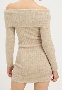 Robe beige à épaules dénudées en tissu maille côtelée, avec manches longues et silhouette ajustée. Texture douce avec un motif subtil.
