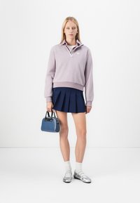 Young woman wearing lavender quarter-zip sweatshirt, navy pleated skirt, white socks, silver flats, holding small blue handbag against white background.
