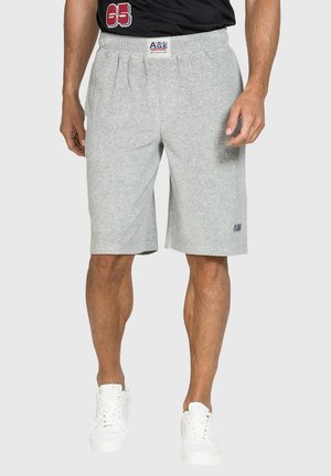 Uomo che indossa pantaloncini grigio chiaro fino al ginocchio con elastico in vita e scarpe da ginnastica bianche, in piedi davanti a uno sfondo semplice.