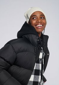 Chaqueta de plumas negra con capucha, que presenta un material brillante y una textura acolchada. Gorro blanco con detalle bordado. Camisa de cuadros en capas por debajo.