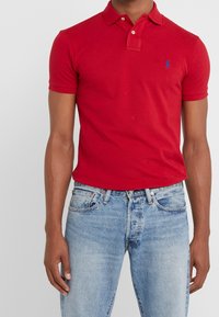 Czerwona bawełniana koszulka polo z klasycznym kołnierzykiem, krótkimi rękawami i niebieskim logo na piersi, zestawiona z jasnoniebieskimi dżinsami.