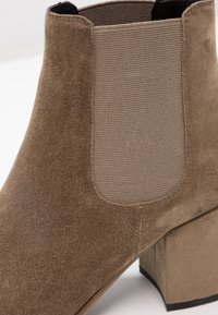 Gros plan d'une bottine en daim taupe avec un panneau latéral élastique et un talon bloc sur fond blanc.