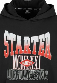 Čierna mikina s kapucňou, ktorá obsahuje odvážny, gradientný biely a červený text. Dizajn zahŕňa slová "STARTER" a "MCMLXXI" spolu s grafickým vzorom hviezdy.