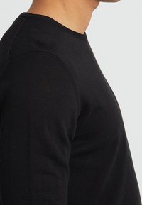 Schwarzes Langarmshirt in taillierter Passform mit einem runden Halsausschnitt und strukturiertem Stoff, von der Seite betrachtet.
