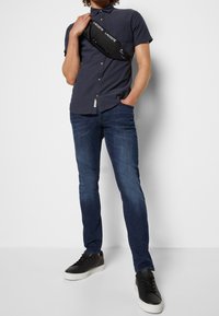 Krátký rukáv, námořnický vzorovaný košile s knoflíky, tmavě modré slim-fit džíny a černé tenisky s bílou podrážkou. Černá taška na rameno nošená přes tělo.