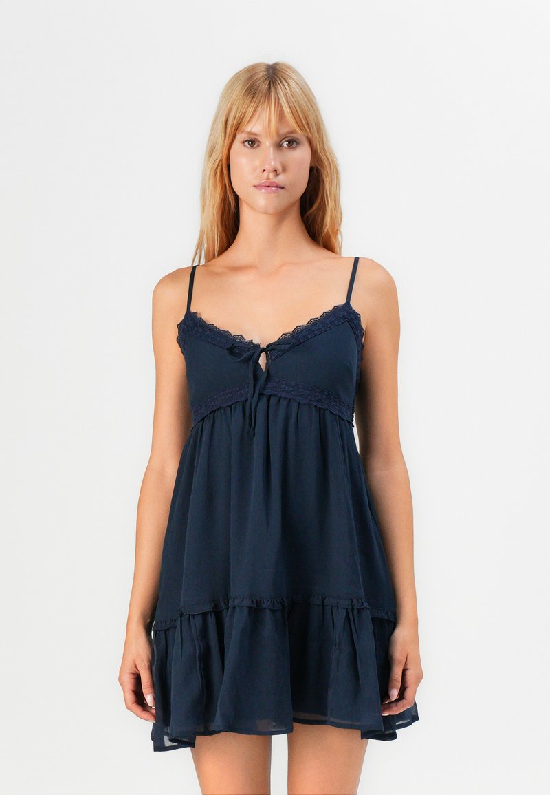 Vrouw in een mouwloze, marineblauwe jurk met kantafwerking, spaghetti-bandjes, een gestrikte details aan de voorkant en een gerimpelde zoom, staand tegen een witte achtergrond.