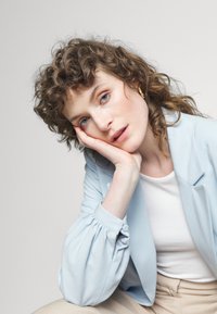 Blazer azul claro com mangas bufantes, usado sobre uma blusa branca. Modelo descansa o queixo na mão, tem cabelo encaracolado e está sentado contra um fundo neutro.