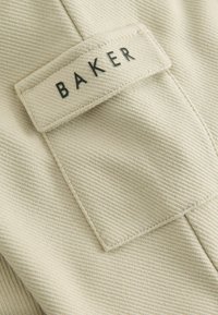 Beige kangasta, jossa on uurrettu pinta. Siinä on suorakulmainen etiketti, jossa lukee "BAKER" mustalla. Yksinkertainen muotoilu, istuva malli.