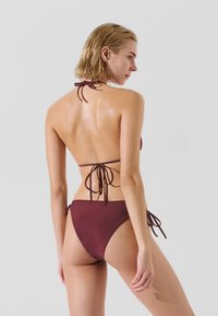 Burgundi színű bikini szett texturált felülettel, nyakpánttal és oldalt megkötős alsó résszel, minimális takarással és állítható mérettel.
