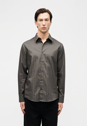 Mees, kellel on seljas pika varrukaga, tumesinine nööpidega särk peene mustrilise kujundusega, seisab tavalise heleda tausta ees.