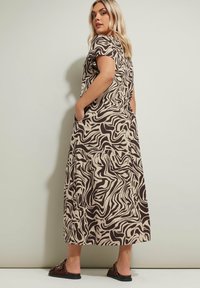 Maximekko vaaleassa värissä, jossa on ruskea abstrakti kuvio, lyhyet hihat, sivutaskut, jokseenkin löysä istuvuus ja helma, joka ulottuu nilkojen alapuolelle.