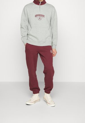 Felpa grigia con collo a metà zip, dettagli rossi e logo Converse; abbinata a pantaloni jogging maroon con dettagli del logo; indossata con sneakers beige.