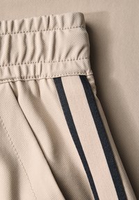 Beige fabric with a textured finish. Elastic waistband and a side panel featuring two thin navy stripes. Neatly stitched seams.