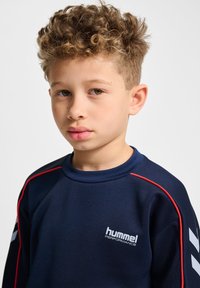 Dreng med krøllet hår iført en marineblå Hummel Performance sweatshirt med røde kantbånd og hvide geværstriber på ærmerne.