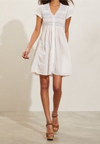 Donna che indossa un vestito bianco a maniche corte con bottoni sul davanti, decorato con ricami blu e sandali con tacco alti marroni, in piedi su un pavimento chiaro.