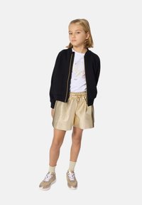 Zwarte bomberjack met gouden rits, witte graphic t-shirt, gouden metallic shorts en lichtgouden sneakers met witte veters.