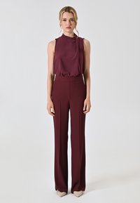 Burgunderfarbene ärmellose Bluse mit gerafftem Detail am Ausschnitt, kombiniert mit passenden weiten Hosen aus einem glatten, fließenden Stoff.