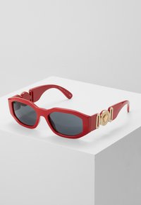 Gafas de sol de plástico rojo con un diseño geométrico, lentes oscuros y detalles en dorado con un acento circular en la montura.