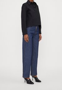 Camicetta nera corta con colletto, abbinata a jeans in denim blu a gamba larga con cuciture visibili. Indossata con scarpe con tacco alto nere.