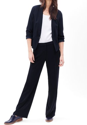 Blazer marine à coupe décontractée et à revers ajustés, associé à un pantalon large assorti et un top blanc, porté avec des chaussures foncées.