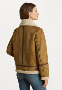 Giacca in suede marrone con colletto in shearling, caratterizzata da dettagli beige, un taglio dritto e cuciture orizzontali lungo la parte posteriore e le maniche.