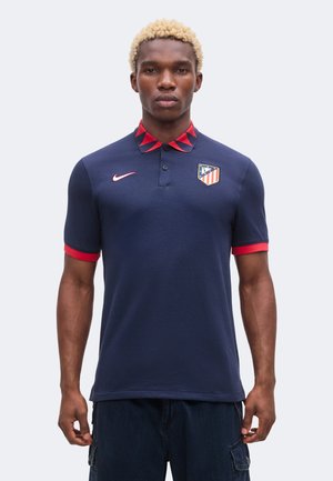 Homme portant un polo bleu marine avec des accents rouges, logo Atlético Madrid sur la poitrine et le logo Nike, debout devant un fond uni.