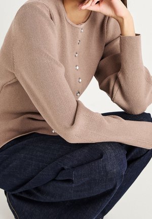 Personne portant un pull beige côtelé avec des boutons sur le devant, reposant son menton sur sa main, et un pantalon bleu foncé visible.