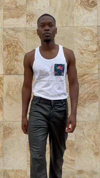 White tank top with floral pocket, paired with black leather pants. Model stands against a textured beige wall.