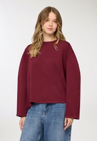 Felpa bordeaux a girocollo con maniche lunghe, vestibilità comoda e superficie texturizzata. Abbinata a jeans ampi azzurri.