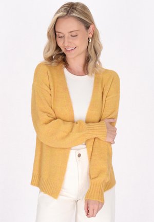 Cardigan giallo a maglia con una vestibilità comoda, apertura frontale, polsini e orlo a coste. Indossato sopra una maglietta bianca e abbinato a pantaloni chiari.